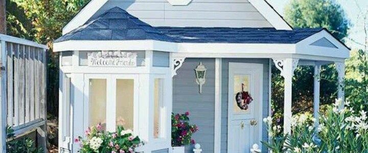 A warm and colorful photo of a tiny blue and white cottage, featuring victiorian and french country styled exterior molding and embellishments. There is a bay window and sheer curtains shine through. Outside is a lovely white rose bush and meticulously detailed landscaping A front porch has a little white bench and a lovely blue and white welcoming wreath on the front door. #CottageStyle, #FrenchCountry, #HomeDecor, #Relaxation, #ComfortableLiving
